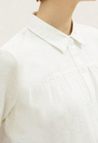 Camicia bianca con colletto realizzata in tessuto texturizzato con un sottile motivo a macchie, dotata di chiusura a bottoni e dettagli plissettati sotto il giogo.