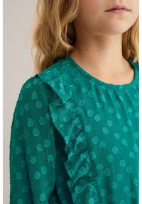 Tirkīza blūze ar apaļu izgriezumu, kas rotāta ar teksturētu punktu rakstu un kroku akcentiem uz pleciem un piedurknēm. Viegls, caurspīdīgs audums.