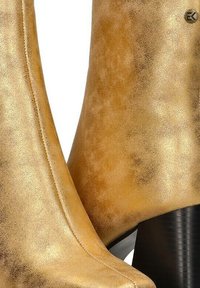 Botines metálicos dorados con una textura suave, un patrón sutil y un tacón negro alto; diseño elegante con detalles de costura.