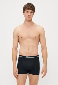 Fekete pamut boxer nadrág, testhezálló kialakítással, márkás elastikus derékkal és sima felülettel. A modell felső nélkül pózol.