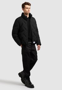 Doudoune noire avec motif en diamant, col haut et fermeture éclair. Portée sur un pull noir, associée à un pantalon cargo noir et des bottes.