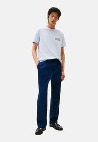 Camiseta de algodón de color azul claro con mangas cortas y un logo en el pecho, combinada con pantalones azul marino. El modelo está de pie con las manos en los bolsillos.