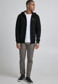 Svart zip-up hettegenser med snøring, kombinert med en hvit T-skjorte, lysegrå bukser og svarte joggesko. Enkelt design og avslappet passform.