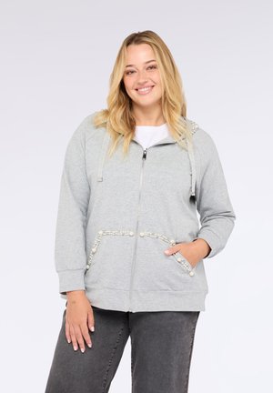 Glunderende vrouw in een lichtgrijze rits hoodie met parel-details op de zakken en capuchon, gecombineerd met donkere grijze jeans, staande met haar hand in de zak.