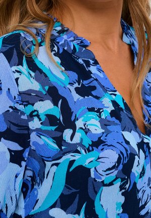 Kvinde iført en blå bluse med blomstermønster i nuancer af turkis, marineblå og lyseblå, der viser halsudskæringen og en del af håret.