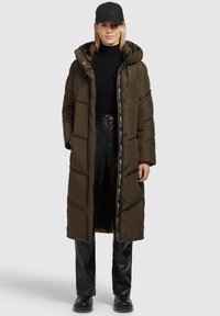 Frau, die einen langen braunen gepolsterten Mantel mit Kapuze, einen schwarzen Rollkragenpullover, schwarze Lederhosen, schwarze Stiefel und eine schwarze Baseballmütze trägt, steht vor einem grauen Hintergrund.