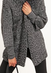 Cardigan oversize texture in nero e grigio, con apertura frontale, maniche larghe e vestibilità comoda. Abbinato a un top grigio a maniche lunghe e leggings.