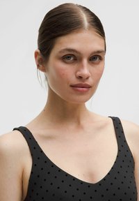 Čierne šaty s bodkami, s výstrihom do V a širokými ramienkami. Hladká textúra, priliehavý dizajn, ktorý zdôrazňuje geometrické tvary.