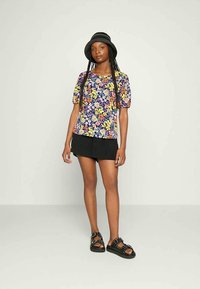 Top de manga curta com padrão floral em roxo, com flores em rosa, amarelo e branco; combinado com calções pretos e sandálias rasas pretas.