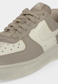 Close-up van beige en witte Nike Air Force 1 sneaker met veters, perforaties op de neus en het zijkant Swoosh-logo.
