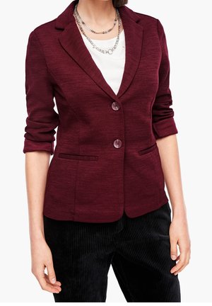 Kobieta ubrana w dopasowaną bordową marynarkę z podwiniętymi rękawami, biały top, wielowarstwowe srebrne naszyjniki i czarne spodnie sztruksowe.