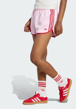 Personne portant un short de sport rose avec des bandes rouges et le logo Adidas, des chaussettes blanches avec des bandes rouges et des baskets Adidas rouges.