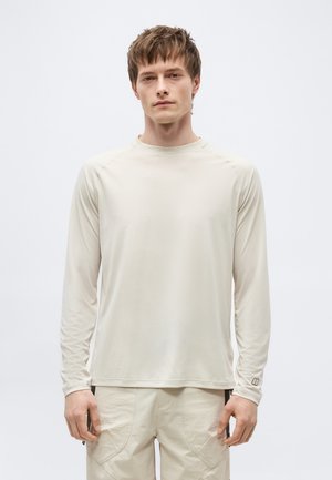 Rövid barna hajú férfi világos bézs hosszú ujjú ingben és texturált bézs nadrágban, egyszínű fehér háttér előtt áll.