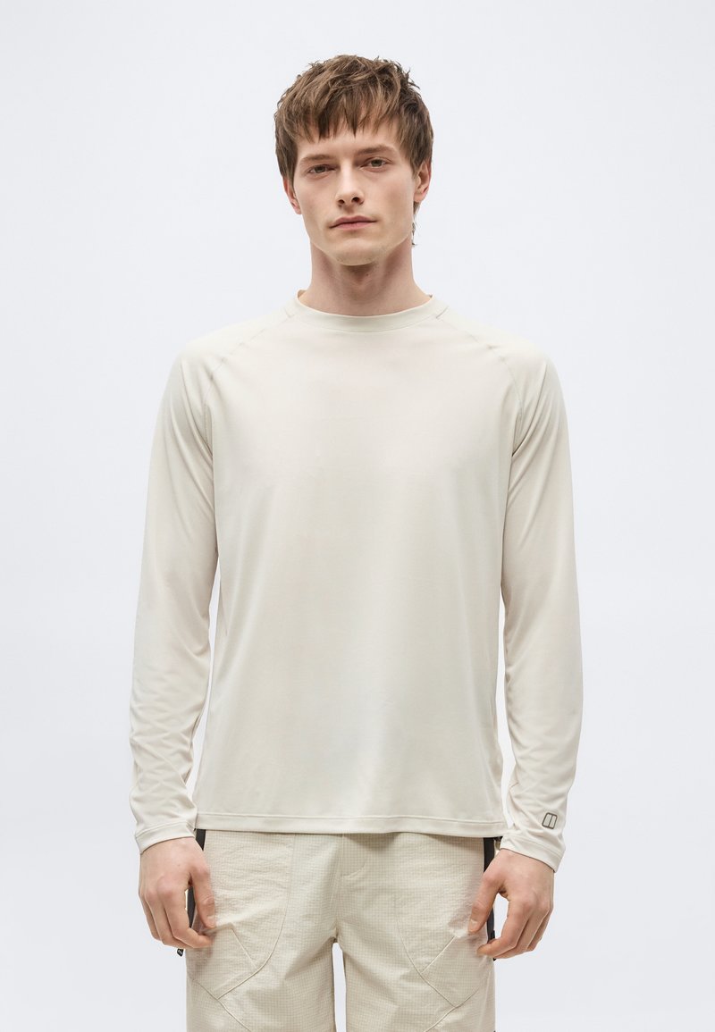 Άνδρας με κοντά καστανά μαλλιά που φοράει ανοιχτό μπεζ μακρυμάνικο πουκάμισο και υφασμάτινο μπεζ παντελόνι, στέκεται μπροστά σε λευκό φόντο.