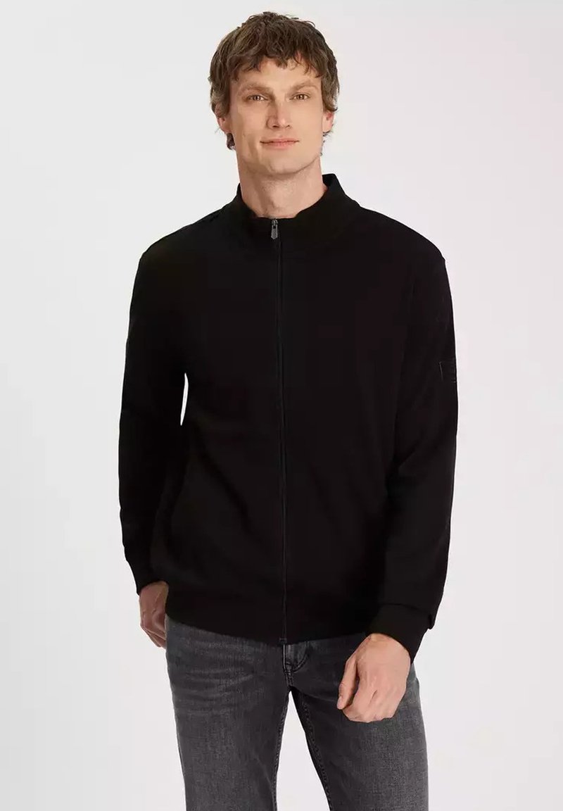 Czarna kurtka z zamkiem na całej długości i wysokim kołnierzem, z prążkowanymi rękawami i gładką teksturą. Noszona z ciemnymi dżinsami, prezentując luźny fason.