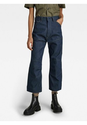 Person trägt dunkelblaue, gekürzte Jeans mit verstärkten Nähten, olivgrünes Hemd, beige Socken und schwarze klobige Stiefeletten.