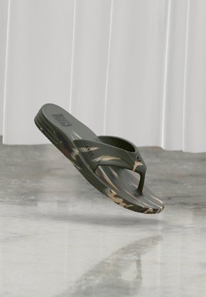 Une sandale tongs unique vert olive et à motif camouflage flottant au-dessus d'un sol en béton poli avec des rideaux blancs en arrière-plan.