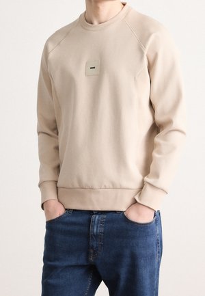Sweat-shirt beige à manches longues avec col rond, petit patch logo "BOSS" sur la poitrine, porté avec un jean bleu, mains dans les poches.