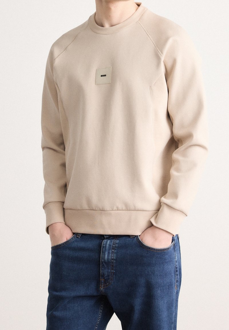 Sweat-shirt beige à manches longues avec col rond, petit patch logo "BOSS" sur la poitrine, porté avec un jean bleu, mains dans les poches.