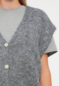 Gray knitted vest with a V-neck, short sleeves, and two large cream buttons. Worn over a light gray short-sleeve T-shirt.