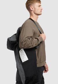 Mladik u smeđem dukserici nosi crnu jaknu s vidljivom bijelom etiketom i drži remen preko ramena, bočni prikaz.