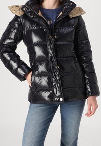 Schwarze Steppjacke mit glänzendem Finish, ausgestattet mit einer khakifarbenen Kapuze, Reißverschluss vorne und Druckknöpfen. Das Design umfasst horizontale Steppnähte.