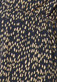 Primo piano di un tessuto scuro con cucitura arricciata e un motivo irregolare di piccole macchie ovali beige e gialle.