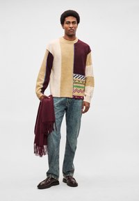 Heegeldatud kampsun mitmesuguste beežide, burgundi ja kreemikaunistuste mustritega; kombineeritud siniste teksade ja burgundi tooni narmastega salliga. Tumedad kingad.