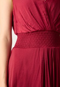 Robe bordeaux avec une taille smockée, présentant un tissu doux et drapé. La partie supérieure a un décolleté profond et une jupe fluide.