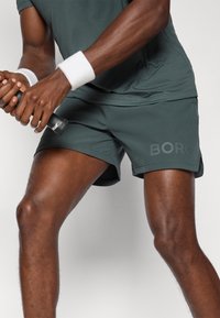 Calções de desporto verde-escuro com um elástico na cintura, apresentando um logótipo "BORO" numa tonalidade mais clara. Textura suave e comprimento até à metade da coxa.