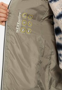 Binnenkant van de jacket van gerecycled stof, licht beige met een zachte textuur. Bevat gele iconen die waterdicht, ademend, winddicht en getapete naden aangeven.