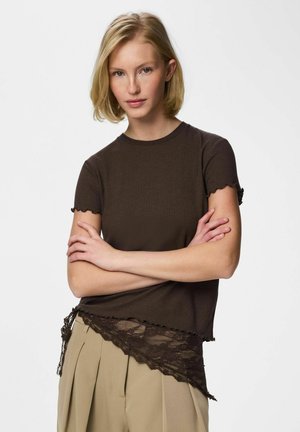 Plavuša s kratkom kosom u tamnoljubičastoj majici kratkih rukava s čipkastim rubom i bež plisiranim hlačama, prekriženih ruku.