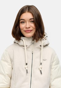 Young woman with shoulder-length brown hair wearing a cream-colored hooded jacket with rope drawstrings and quilted sleeves, smiling gently.