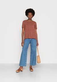 Top de mangas curtas castanho com padrão, jeans de perna larga azul claro, tamancos castanhos, segurando uma bolsa de palha. Textura suave, ajuste casual e design descontraído.
