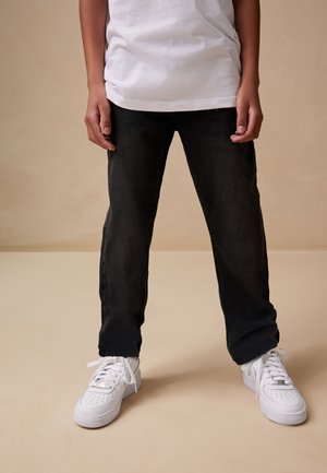 Barn iført sorte jeans, hvide sneakers og en hvid top står mod en ensfarvet beige baggrund.