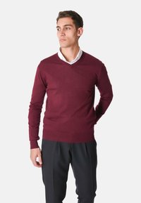 Maglione a maglia bordeaux con scollo a V, design aderente, polsini e orlo a coste, abbinato a pantaloni neri, indossato sopra una camicia bianca.