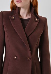 Bruine wollen mantel met dubbele knopenrij en een hoge kraag, voorzien van goudkleurige knopen in bloemvorm en een gladde, gestructureerde stof.