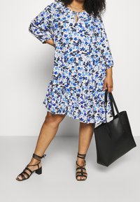 Bloemenjurk in blauw en zwart tinten, driekwart mouwen, sleufhals, gelaagde zoom, gecombineerd met zwarte sandalen en een bijpassende tote bag.