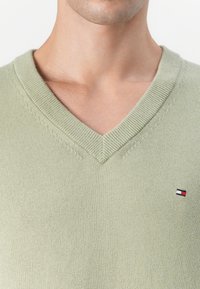 Lys grønn v-hals genser laget av strikket stoff, med ribbestrikket krage og en liten logo-lapp på brystet. Glatt tekstur og slim-fit design.