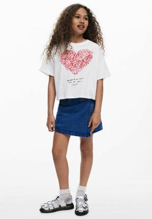 Witte cropped t-shirt met een rood hart afbeelding en tekst; gecombineerd met een denim rok en zilveren sandalen, met witte kanten sokken.