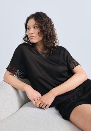 Νεαρή γυναίκα με σγουρά μαλλιά που χαλαρώνει σε ένα ανοιχτόγκρι καναπέ, φορώντας μαύρο τοπ με δαντέλα και μαύρο σορτς που ταιριάζει.