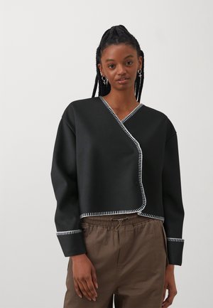 Zwarte cropped wrap top gemaakt van een glad materiaal met witte stikselaccenten aan de randen. Lange mouwen en een V-hals ontwerp.