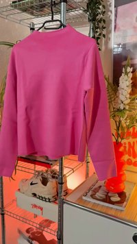 Top roz cu mânecă lungă și guler înalt, realizat dintr-un material neted, având o croială lejeră și slituri laterale la tiv.