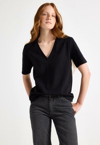 Czarny t-shirt z dekoltem w serek wykonany z bawełny, z krótkimi rękawami i luźnym krojem. Noszony z ciemnoszarymi dżinsami, na jednolitym tle.