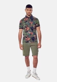 Polo shirt met een tropische bloemenprint in groen, roze en blauw, gecombineerd met olijfgroene cargoshorts en witte sportschoenen.