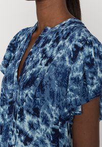 Blůza s modrým batikovaným vzorem vyrobená z lehkého materiálu, se sklady na přední části a krátkými volánkovými rukávy. Vzor zobrazuje různé odstíny modré.