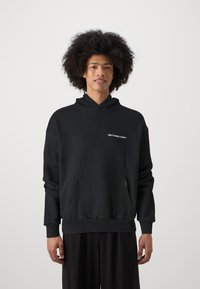 Schwarzer Kapuzenpullover mit einer Vordertasche, mit weißem Text "don't waste culture" auf der oberen linken Seite. Weicher Stoff mit lockerer Passform.