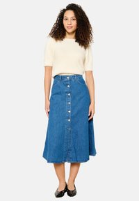 Denim krilo z visokim pasom, oprijetim zgornjim delom, A-linijsko obliko in sprednjim gumbom. V družbi kratkotrajnega kremnega puloverja.