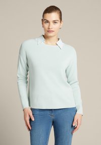 Helles mintgrünes Sweater mit rundem Halsausschnitt und kontrastierendem weißen Kragen. Weiche Textur, lange Ärmel und lässige Passform. Kombiniert mit blauen Jeans.