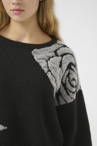 Woman wearing a black sweater with a large gray rose pattern on the shoulder against a plain background.